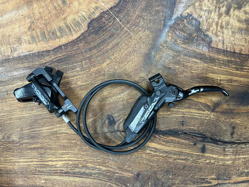 A black SRAM hydraulic disc brake lever and caliper assembly with a coiled black cable, displayed on a rustic wooden surface.