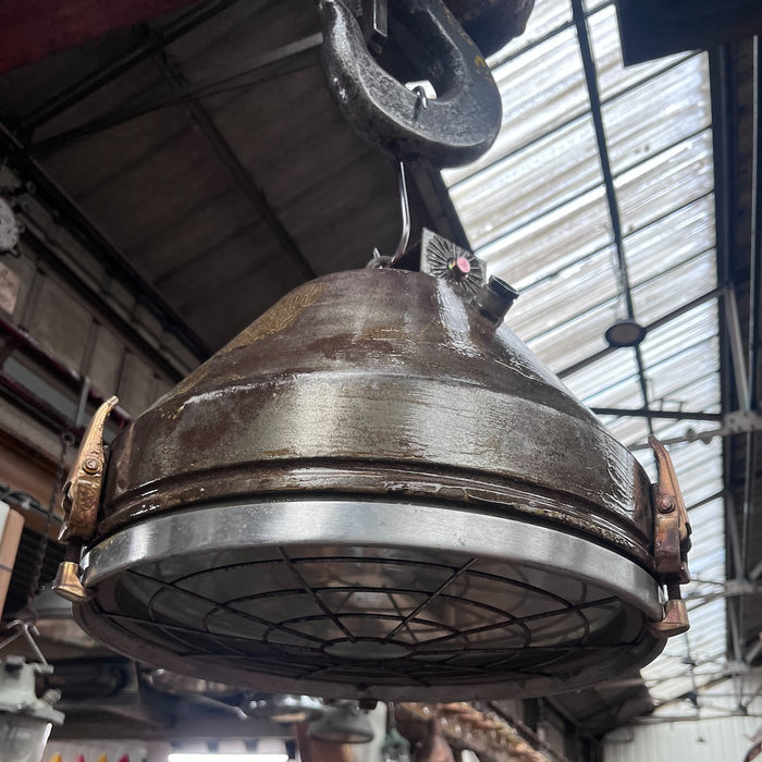 Salvaged VEB industrial steel ceiling light hanging from a hook, showing conical body, brass clamps and wire-guarded glass front
