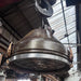 Salvaged VEB industrial steel ceiling light hanging from a hook, showing conical body, brass clamps and wire-guarded glass front